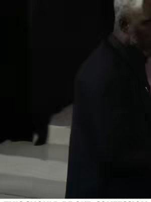 A man with white hair in a dark setting is walking. The lighting is dim and the image is a medium shot capturing the man from the waist up. The man is wearing a dark jacket or blazer and the background is dark.