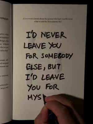 An open book displays handwritten text. The text, written in black marker on the page, starts with "I'D NEVER LEAVE YOU." Words are vertically stacked, with a few teardrop stains visible on the page.