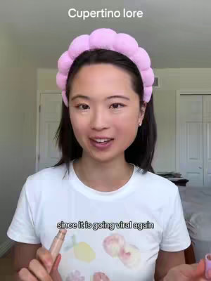 A young woman with dark hair and a light complexion is centered in the frame, wearing a white t-shirt with a colorful print. She is wearing a pink padded headband and appears to be applying makeup with a makeup product in her left hand. The background features two closed white doors and visible ceiling lights. Text overlays include "Cupertino lore" at the top and "since it is going viral again" at the bottom.