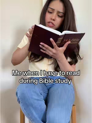 A young woman with long, dark hair is seated on a stool, holding an open book labeled "Holy Bible." She appears to be reading intently with a look of slight exasperation. The background is a simple, light-colored wall.