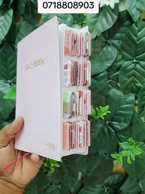 A hand holds a light pink Bible with multiple tabs visible. The tabs are labeled with the names of books of the bible. It is set against a backdrop of green foliage.