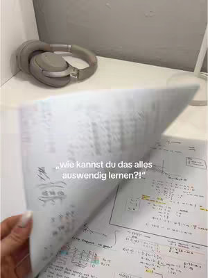 A hand flips through pages of notes covered in formulas and diagrams. The page is partially obscuring the view, revealing some study materials. In the background, a pair of headphones sits on a shelf.