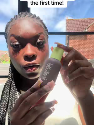 A close-up shot featuring a woman holding a skin tint product in front of her face. The lighting appears natural, with the background suggesting it's outdoors. The woman has long braids, and is smiling slightly with her mouth open.