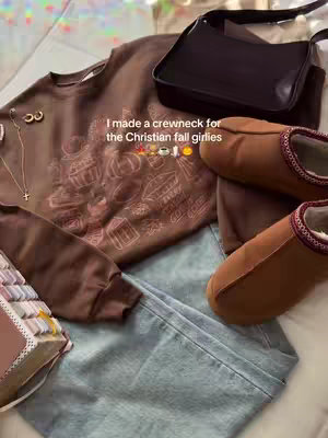 The image is a flat lay featuring various fall-themed items arranged on a light blue surface. At the center is a brown crewneck sweatshirt with a fall-themed design. Surrounding the sweatshirt are complementary items: a black purse with a long strap, a pair of brown plush slippers, blue jeans, a small gold necklace with a cross, and the corner of an open bible. The composition is warm-toned, emphasizing browns and creams.