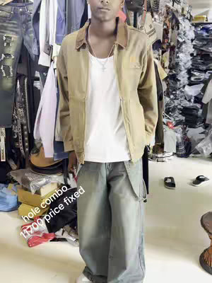 A young man stands indoors, posed in front of a cluttered backdrop of clothes and accessories. He's wearing a tan jacket over a white t-shirt, paired with light blue, loose-fitting jeans. The image is brightly lit, and the man gazes directly at the camera, with added text in the bottom left corner.