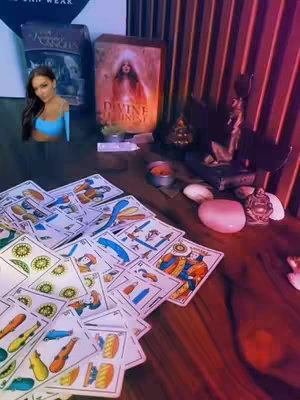 A wooden table is scattered with tarot cards, with a few fanned out in the foreground. A box labeled "Divine Destiny" with a picture of a person is visible in the background, alongside other items like candles. The overall lighting has a deep red hue.