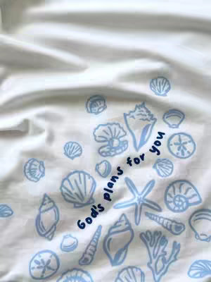 A white fabric with numerous light blue seashell illustrations and the text "God's plans for you" in a handwritten font. The composition is organic, with the shells scattered across the fabric.