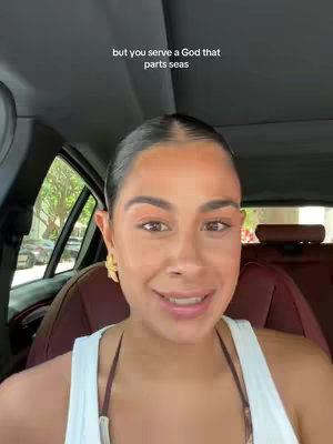 A woman is filmed from the chest up, inside a car. She is smiling and looking at the camera. The background shows the inside of the car and some partially visible street scenery.