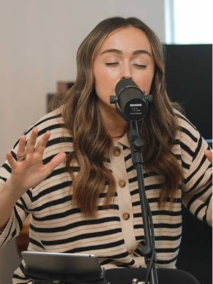 A woman with long, wavy brown hair is singing into a microphone. She is wearing a striped shirt and has her eyes closed. Her hands are raised in the air.
