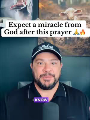 A man wearing a black shirt and a baseball cap that says 'With God' in white letters is smiling at the camera. Behind him are two framed images; one shows a lamb in a field, and the other depicts a person's hands. Text on the upper left says 'Expect a miracle from God after this prayer'.