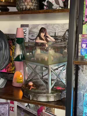 The image showcases a collection of manga and anime-related items. The focal point is a Tomie Kawakami statue by Echoes Gallery, displayed within a clear container filled with a blueish liquid, sitting on a metal structure. Various backgrounds with drawings/paintings are partially visible. The scene is set on a shelf that includes framed artwork, a lava lamp, and other items, indicating a collector's display.