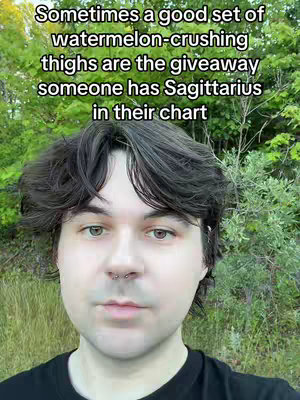 A person with short, dark hair and light eyes gazes directly at the camera. The background features a blurred view of green foliage. Text overlays on the image.