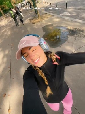 A selfie of a woman in a cap and headphones smiling brightly while walking outdoors, possibly in a park or city environment.  The background is blurred, showing trees and other people, while the focus is on her joyful expression.