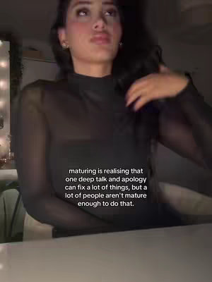 A woman with dark hair is featured in a dim, indoor setting. She is wearing a black, sheer-sleeved top. The background is blurred. The text overlay, written in white sans-serif font, presents a philosophical statement about maturity and communication.