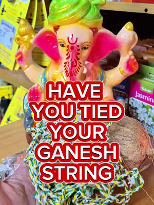 The image showcases a colorful, cartoonish statue of the Hindu deity Ganesha, adorned in bright hues of yellow, pink, and green. The statue is the focal point, with a red-outlined text overlay that poses a question concerning a "Ganesh string." The background is a soft blur of items that suggest a store or marketplace setting.