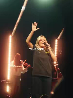 The image is a stage performance scene. A woman with blonde hair, wearing a dark shirt and black pants, is singing into a microphone. She has her right arm raised, and there's a background of stage lighting. In the background, a violinist is playing on a stage.