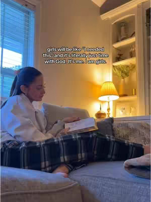 A woman is sitting on a gray couch and reading a book; the room has a window, a lamp and a white cabinet.