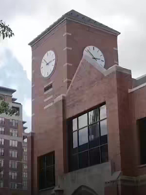 The image shows a brick clock tower against a cloudy sky. Visible clock faces on the tower display the time. Some windows are visible on the tower. The building next to the tower has other windows.
