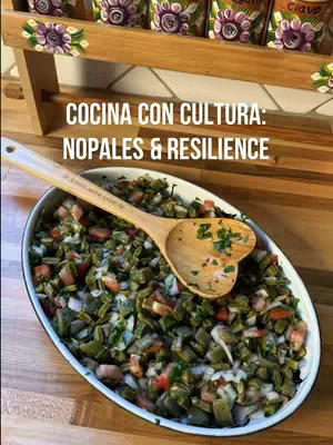 A top-down view showcases a white oval dish filled with a vibrant salad, a wooden spoon with a heart-shaped end resting on it. The salad features a colorful mix of green nopales (cactus), red tomatoes, and white onions, all finely chopped. The background includes a wooden surface and a wooden rack with small decorative jars.