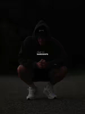 A person wearing a black hoodie is crouching in a dark setting. The person's face is obscured by the hood, and only the lower body and white sneakers are visible. A caption 'HARDSHIPS' appears over the chest.