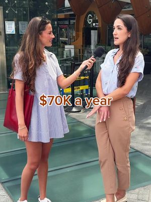 Two women are outdoors; one holds a microphone and interviews the other. The interviewee gestures with her right hand. The overlay text displays the annual salary. The background is a blurred cityscape with various buildings and signs.