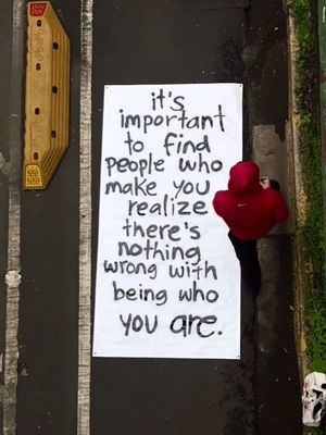 An overhead shot of a person standing on a paved surface, holding a large white sign. The sign has black text on it, and the person is wearing a hooded red sweatshirt. A yellow rectangular object with black dots is visible on the left, and a skull statue is on the bottom right.