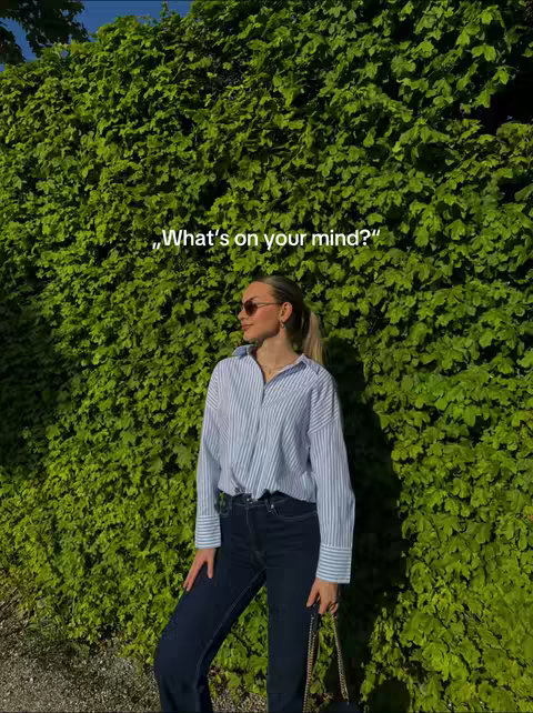A woman wearing sunglasses, a striped button-down shirt, and dark jeans stands in front of a wall of green foliage. The text overlay says, "What's on your mind?"