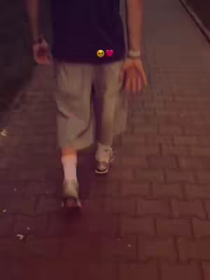 The image features a person walking down a brick path at dusk or night. The person is seen from the waist down, with their back to the camera. They are wearing light gray shorts, a dark shirt, and white socks with white sneakers. The brick path appears to be lit by streetlights.