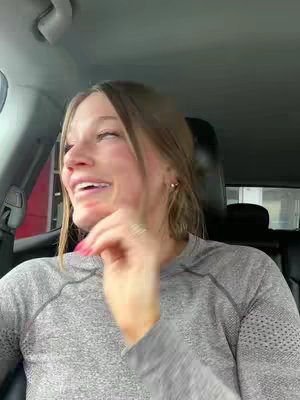 A woman in a car is smiling, with her head turned to her left. Her left hand is raised, and her fingers are near her cheek. She appears to be talking and is wearing a gray long-sleeved shirt.
