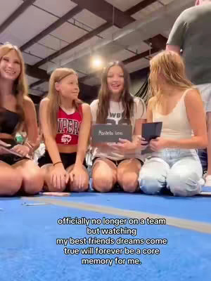 The image shows four young women kneeling close together on a blue floor in what appears to be an indoor sports facility. The women are smiling and looking towards the camera. One of them is holding a sign that reads “Mariella Lem” in block letters. One is holding a small black item, and another is holding a cell phone. The background includes sections of a gym, with lights and a ceiling visible in the distance, and a man is seen standing toward the right of the frame.