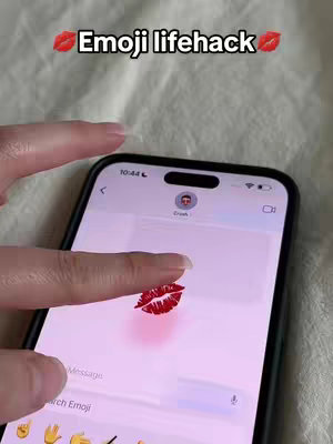 The image shows a close-up of a smartphone displaying a chat interface, with a finger pointing towards a large red lipstick kiss emoji. The phone is held against a soft, light-colored background. The upper portion of the screen displays the time and battery status, while the lower section shows a suggestion to search for emojis. The overlay text at the top says "Emoji lifehack".