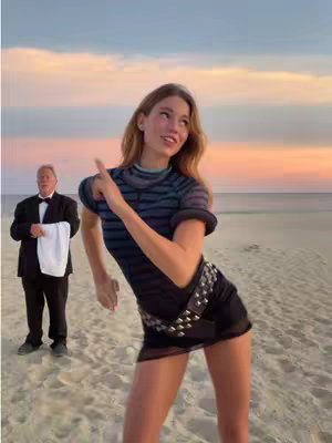 A woman in a black outfit dances on a beach, with a male figure in the background holding a cloth. The color palette is soft, with a sunset background.