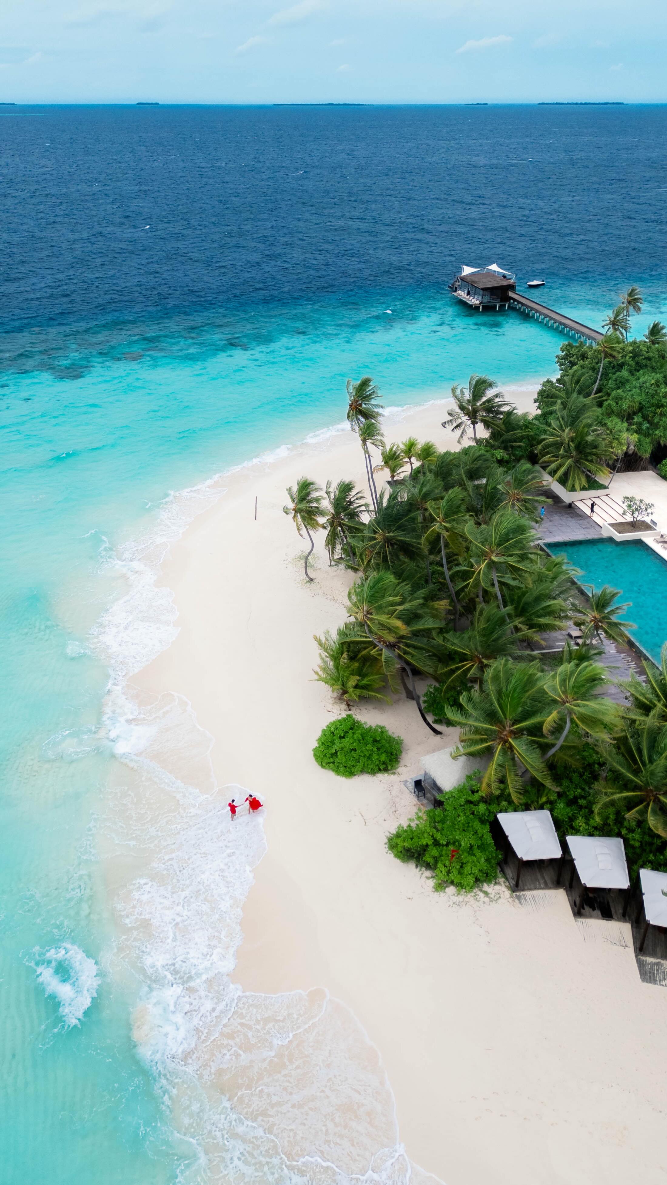 An aerial view of a tropical beach paradise, with crystal-clear turquoise waters meeting a pristine white sand beach. Lush green palm trees line the shore, with a glimpse of a wooden pier extending into the ocean. Two figures in red are walking on the beach. The composition is symmetrical and balanced, showcasing a luxurious resort setting.