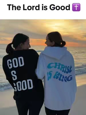 Two women are seen from the back, embracing. One wears a black sweatshirt with "GOD IS GOOD" printed in white, and the other wears a white hoodie that says "CHRIST IS KING" in blue. They appear to be on a beach, facing the sunset.