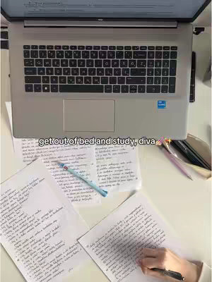 An overhead shot captures a study setup with a laptop, papers covered in handwritten notes, and a hand holding a pen, all on a white surface. The laptop sits at the top of the frame, open and displaying a screen with text. Several sheets of paper with handwritten text are scattered below the laptop, along with pens and pencils. A hand is visible, holding a pen and writing on one of the sheets, suggesting active studying. The composition is clean with cool colors and natural lighting.