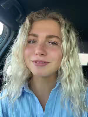 A woman with long, wavy blonde hair is sitting in a car. She is wearing a blue button-down shirt. The image is a close-up, focusing on her face. The background is the inside of the car.