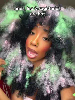 A selfie-style image featuring a woman with colorful curly hair. The hair is predominantly black with sections of green and pink. She has a nose ring and is wearing makeup, including blush. Her mouth is open as if speaking or singing, and text overlays the top reading 'aries, leo & sagittarius are hot'.