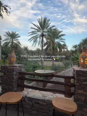 An outdoor cafe with a view of palm trees and a blue sky. There are stone pillars and wooden railings, with stools in the foreground. The text overlay reads 'POV: You found the most peaceful cafe surrounded by nature'.