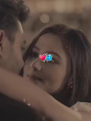 A close-up shot captures a soft, romantic embrace between a man and a woman. Their faces are close, almost touching, suggesting intimacy. A red and blue heart emoji is digitally placed, partially obscuring their faces.. The background is blurred, seemingly lit by warm, soft lights creating a dreamy atmosphere.