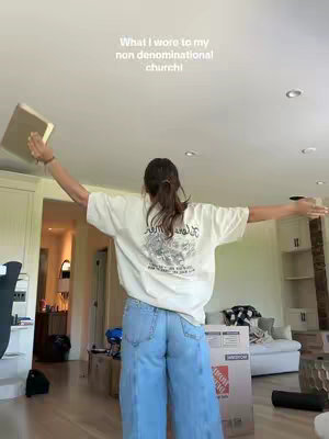 A person is standing with arms outstretched, holding a book in front of them. They are wearing a white t-shirt, blue jeans, and their back is facing the camera. The text overlay says 'What I wore to my non denominational church!'. The background shows a modern interior setting.