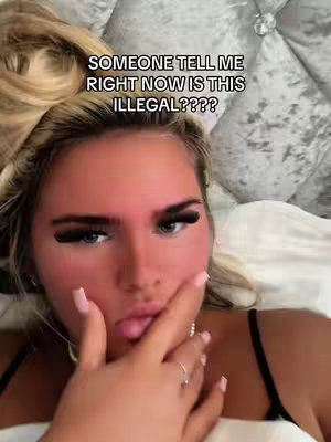 A close-up shot of a young woman's face. She has long blonde hair, large eyelashes, and is wearing light makeup. Her right hand is covering her mouth, and she appears to be looking directly at the camera with a slightly surprised or concerned expression.
