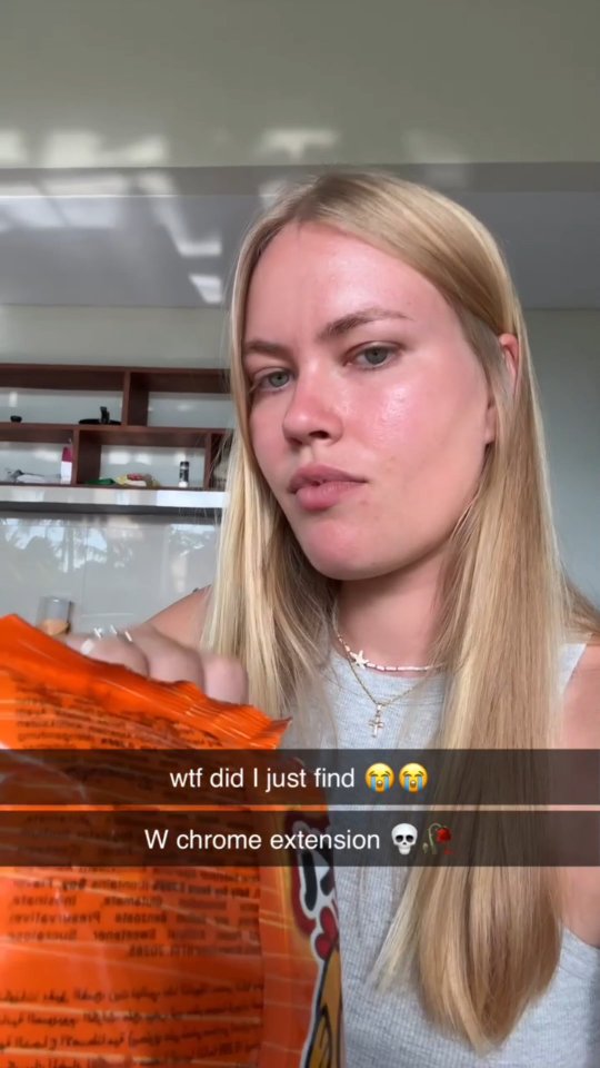 A young woman with blonde hair holds an orange snack bag, with text overlays in a black box at the bottom. The image is a selfie, the woman is in focus, with a plain background. She has a surprised expression. Behind her, there is a hint of what looks like a kitchen.