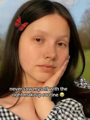 A young woman with dark hair and a butterfly hair clip looks at the camera while touching her cheek. Text on the image says, 'never saw myself with the right makeup routine'. The background has a blurred park scene and a rainbow.