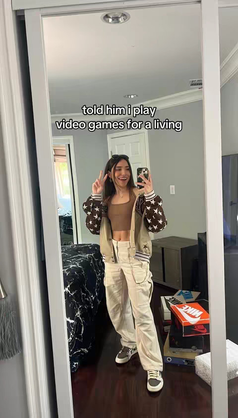 A woman is shown in a mirror reflection, taking a selfie. She is wearing a bomber jacket with star-shaped patterns, a crop top, cargo pants, and sneakers. She is giving a peace sign with her fingers. The background includes a door.