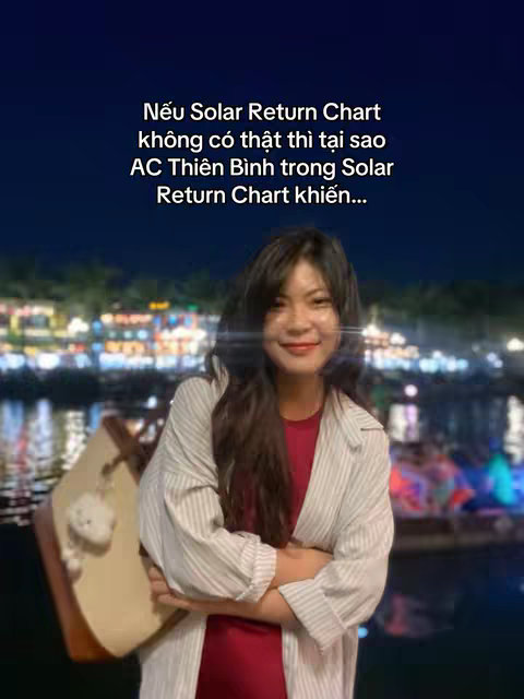 A woman with long, dark hair is the central focus, positioned in front of a blurred background of city lights and water. She's wearing a striped jacket over a red top. The image has overlaid text in the top-left of the image and she is holding an item.
