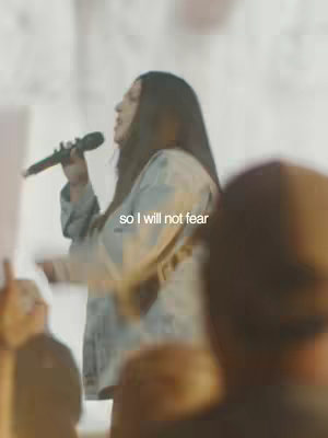 A person, presumably a speaker at a conference, is seen holding a microphone and speaking. The setting is a stage with a blurred background. Text overlayed indicates a message about not fearing.