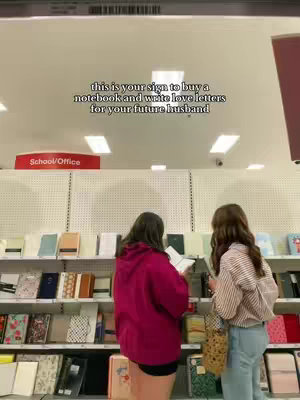 Two women browse notebooks inside a store with white shelves. Overhead, text reads, 'this is your sign to buy a notebook and write love letters for your future husband.'