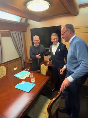 Three men in an office setting, possibly on a train or airplane. They are casually dressed, with one smiling and gesturing towards the others. The room has wood paneling and a large table.