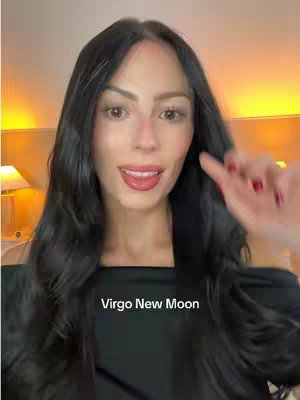 A woman with long, dark wavy hair is smiling, gesturing with her right hand, and partially obscuring the video's background, with a text overlay at the bottom that reads 'Virgo New Moon'. The image has warm lighting.