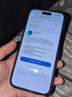 A hand holding a black iPhone displaying a software update screen for iOS 26.1 Beta. The screen shows information about the beta program and prompts to update. The background is a dark, blurry setting suggesting an indoor environment.